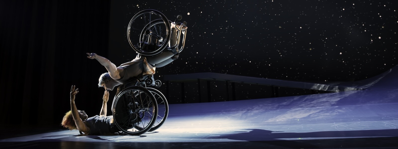 “DESCENT,” Kinetic Light; Laurel Lawson, a white person, balances above Alice Sheppard with arms spread wide, wheels spinning.  Alice, a multiracial Black woman with coffee-colored skin, opens her arms wide to receive her in an embrace. A starry sky fills the background, and moonlight glints off their wheelchair rims. Photo Jay Newman/BRITT.