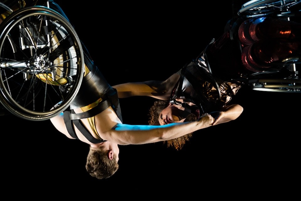 “WIRED,” Kinetic Light; A close-up of Alice Sheppard and Laurel Lawson suspended in the air, arms outstretched and clasping each other’s hands. Alice is a multiracial Black woman with coffee-colored skin and short curly hair; she wears a shimmery deep red costume. Laurel is a white person with cropped hair; she wears a shimmery gold costume with thick black shoulder straps. The dancers are somehow upside down and horizontal at the same time, their wheels shining and facing out; if they let go, they will swing like pendulums. Photo Robbie Sweeny.