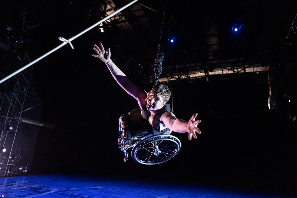 “WIRED,” Kinetic Light; Laurel Lawson, a white person with cropped hair, bounds toward the camera: wheelchair high off the ground, arms open and reaching. A mask of delicate gold wire and lustrous pearls covers half her face. The stage floor below her is drenched in blue light; the same light gives her pale skin a light violet glow. A strand of silver barbed wire appears, close up, in the upper corner. Photo Robbie Sweeny.