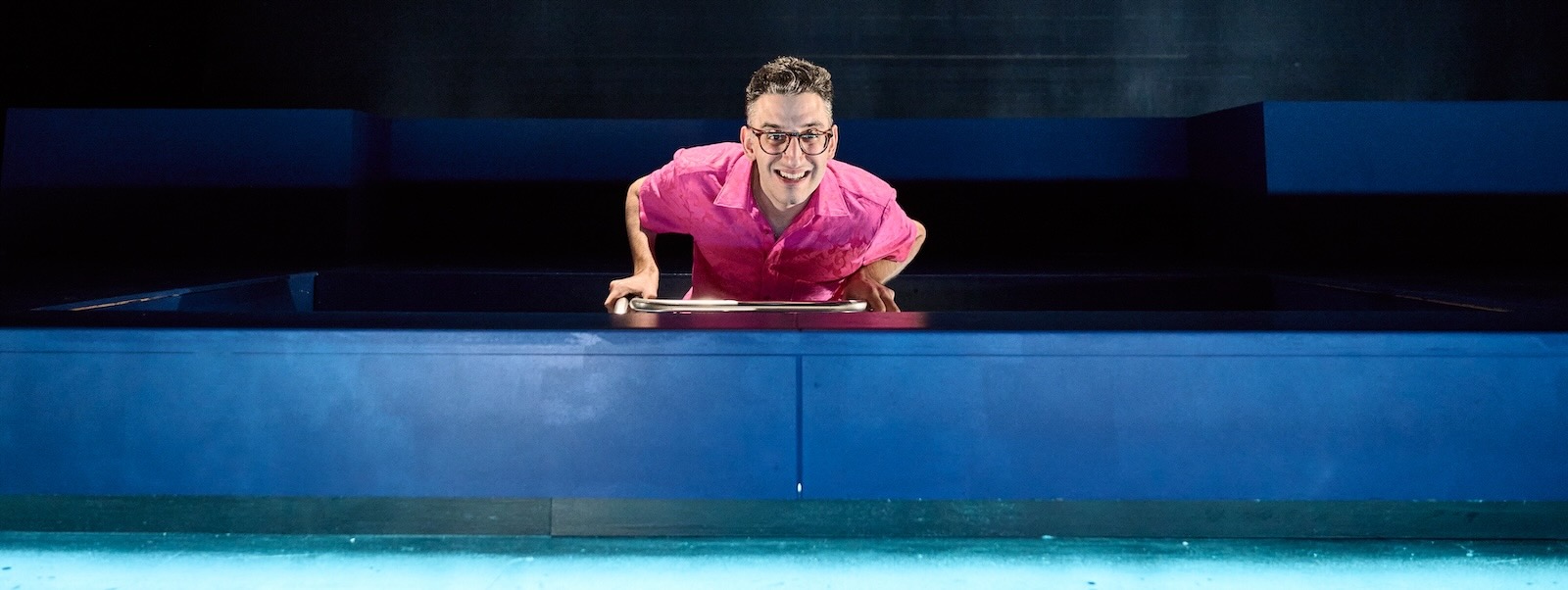 A man with short dark hair wearing a pink shirt leans over a pool.