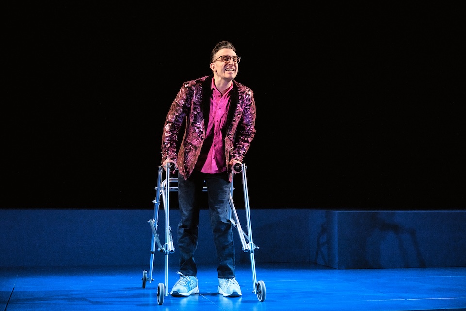 A young man wearing a pink shirt and jacket onstage using a walker.