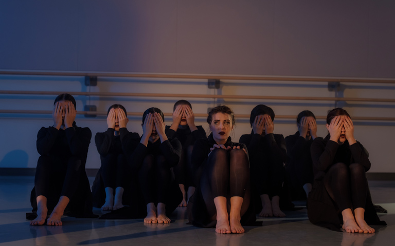 All dancers sit on the floor shielding their eyes except one dancer.