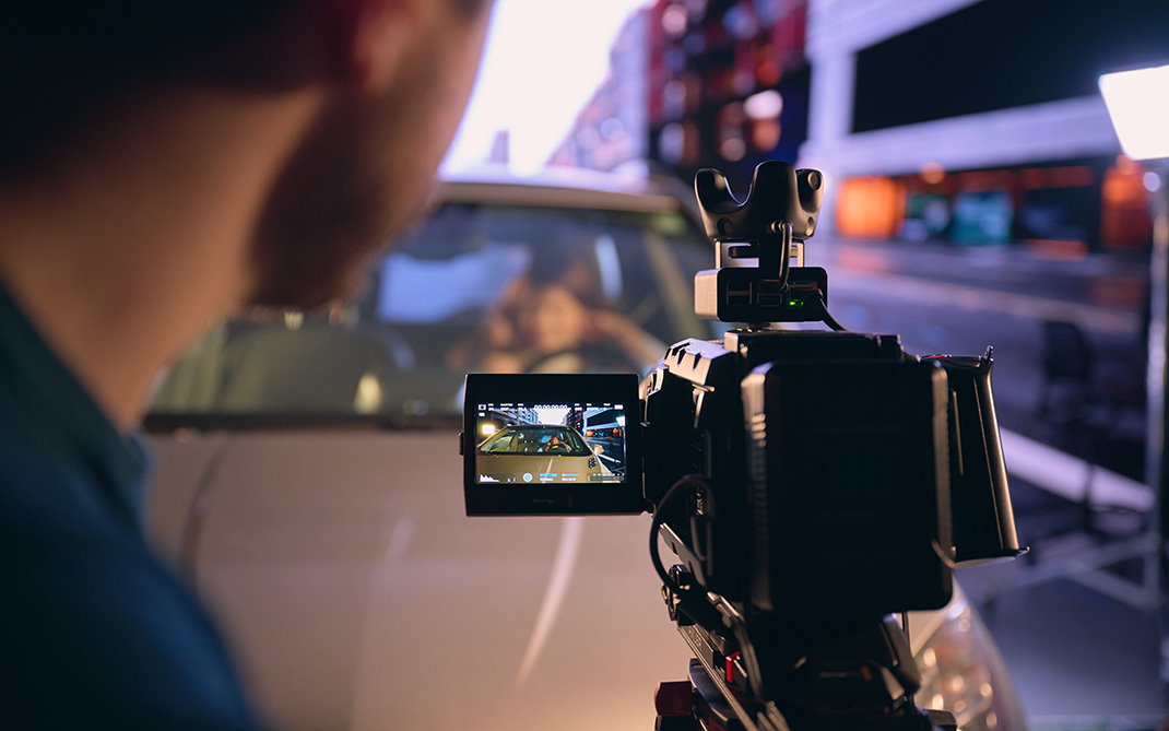 Close-up of a professional camera filming a scene inside a car, with the monitor displaying a clear image of the driver and a blurred studio background.